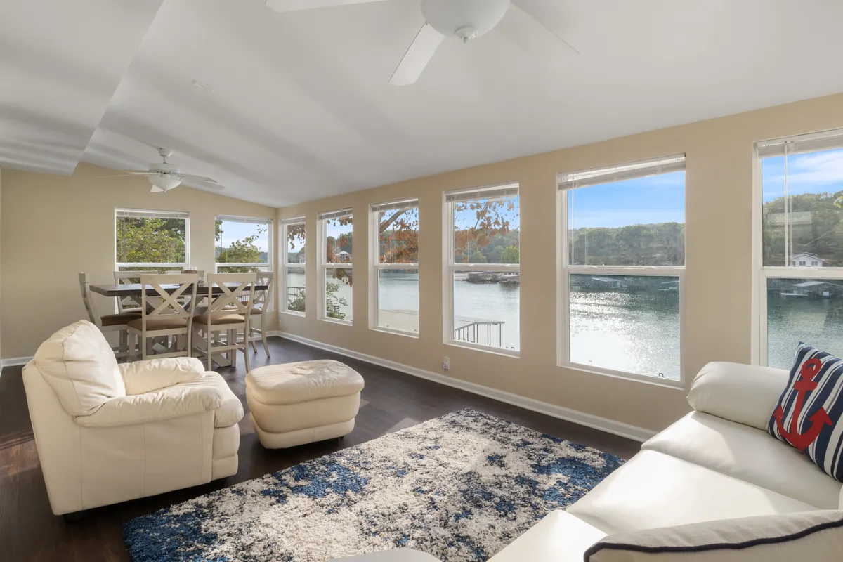 This image features a spacious living and dining area with large windows overlooking the water. Comfortable seating is arranged to encourage relaxation and conversation, while the dining table is set for group gatherings. The natural light streaming in and prominent lake views are central to the scene.