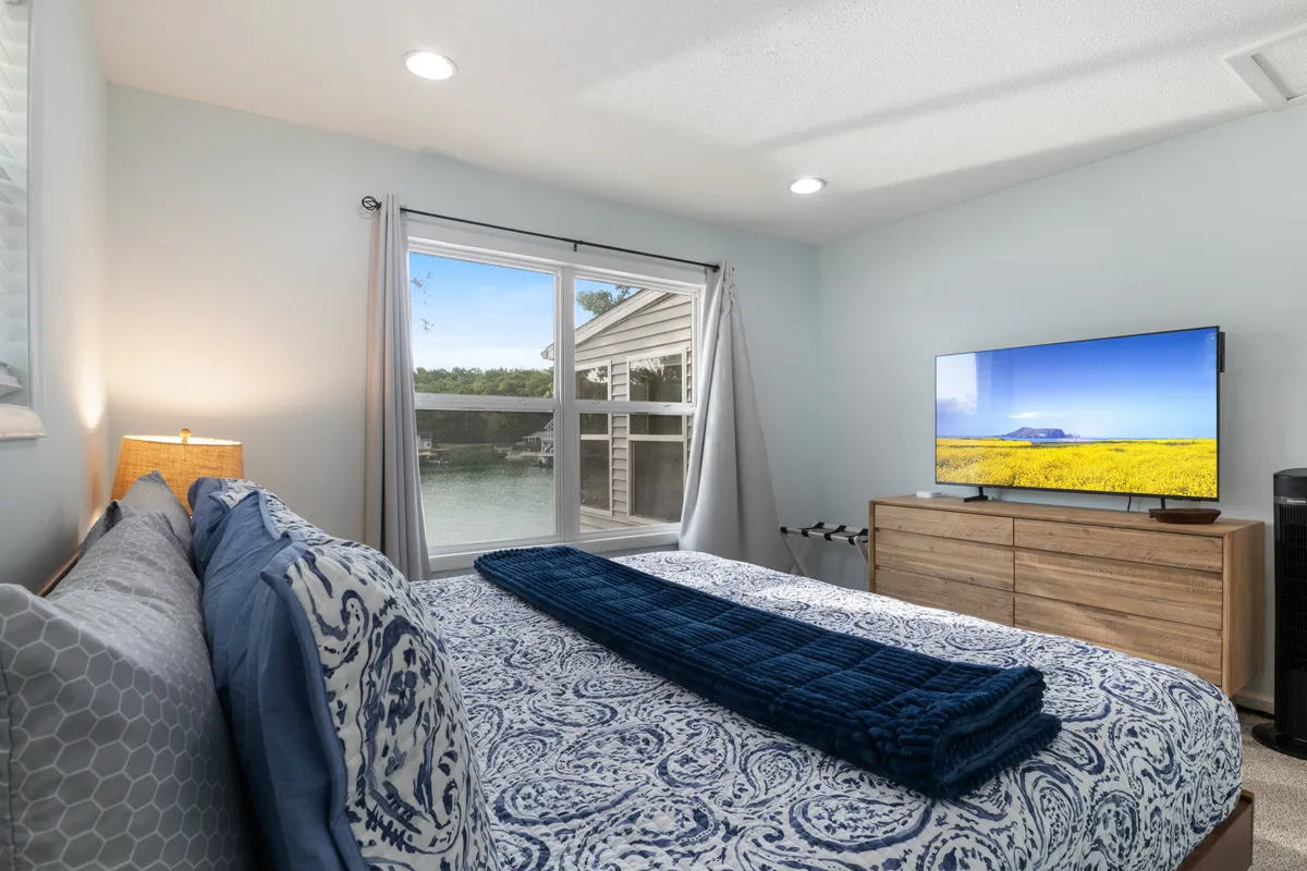 A peaceful bedroom featuring a plush bed with decorative pillows positioned next to a large window that frames a serene view of the water. Soft lighting and a television enhance the sense of comfort and relaxation.