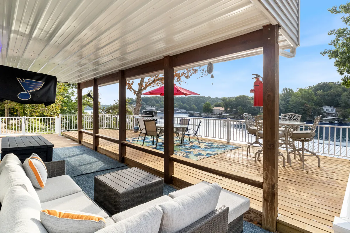 An inviting outdoor seating area featuring a dining table with an umbrella, comfortable chairs, a decorative rug, and an open view of the lake. The expansive deck is positioned for social gatherings and lakeside meals, with plenty of space for kids and adults to relax and play together.
