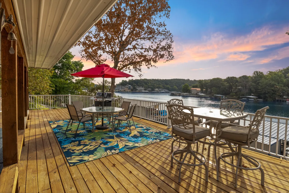 The image showcases an inviting outdoor dining area on a deck overlooking a tranquil lake. The space features a welcoming dining table topped with an umbrella, a colorful area rug that adds a touch of personality, and is surrounded by lush trees with the serene water view as the backdrop. The overall mood is relaxing and ideal for unwinding, socializing, or enjoying the picturesque sunset.
