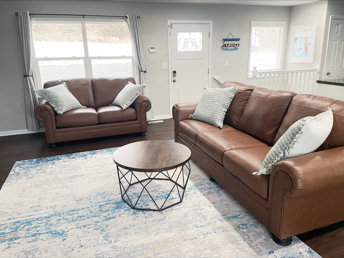A cozy living space featuring brown leather sofas, a round coffee table, a textured area rug, and large windows letting in abundant natural light. This area is clearly designed for family relaxation, entertaining, and intimate gatherings.