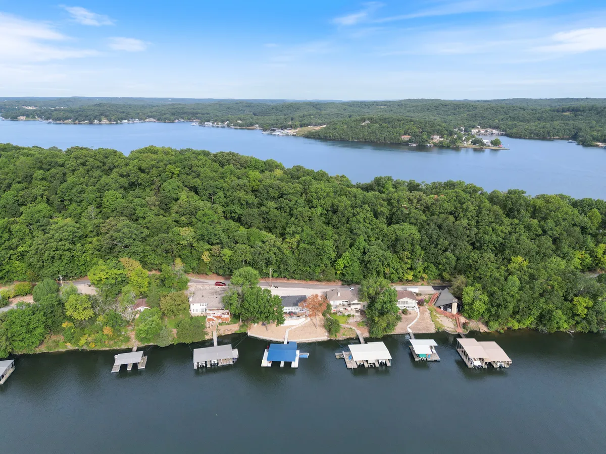 An expansive exterior shot featuring the lake, multiple docks, lush greenery, and homes set back from the shore. The scene evokes a peaceful, natural setting ideal for outdoor activities.
