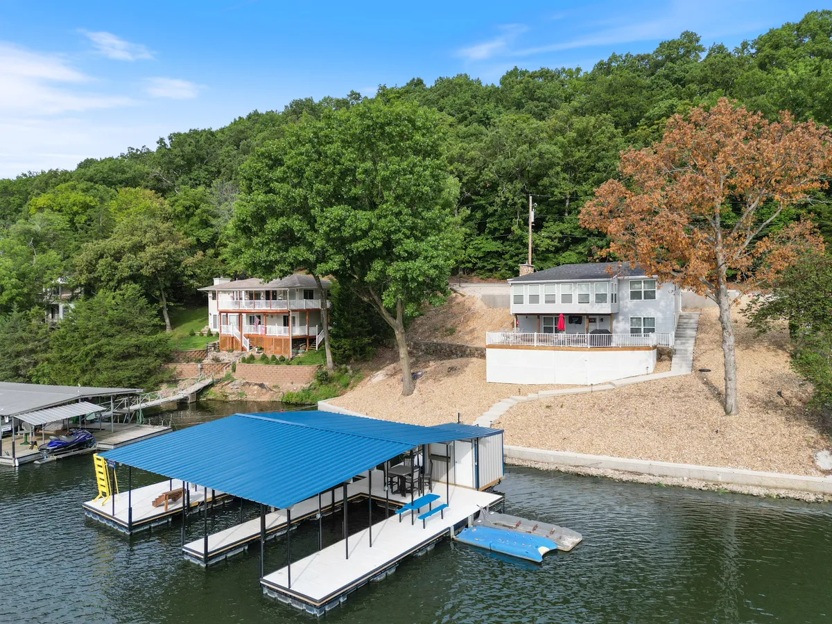 A wide shot of the dock with a blue roof stretching into the lake, surrounded by lush greenery and hillside houses. The calm lake surface reflects the natural beauty, offering a sense of peaceful seclusion. The image suggests opportunities for boating, fishing, and quiet contemplation.