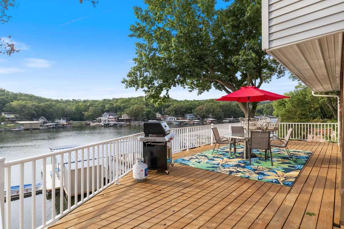 An exterior scene of a grill and outdoor dining table with a red umbrella and decorative rug, set against the backdrop of sparkling lake water. The setup invites guests to enjoy outdoor dining and leisure, framed by the vibrant colors and water views.