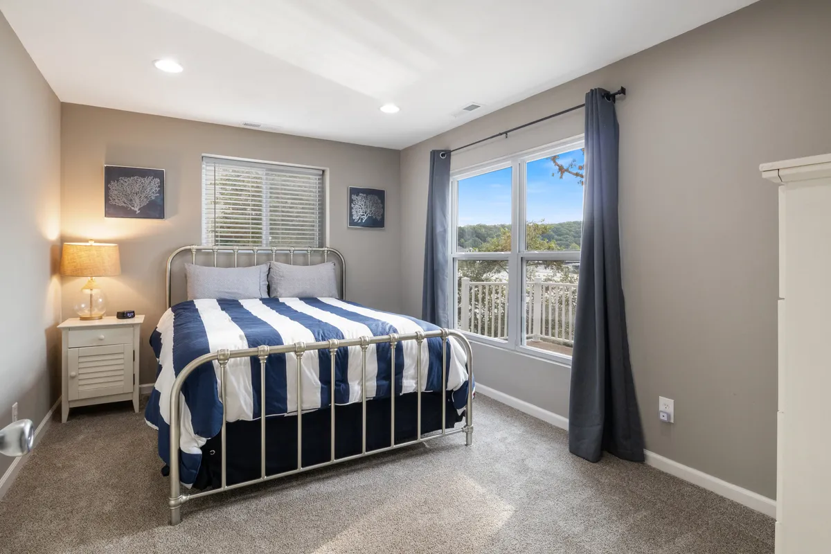 A cozy bedroom with a striped bedspread, nightstand with lamp, and a window letting in gentle natural light, offering views of the outdoors. The room feels peaceful and welcoming.