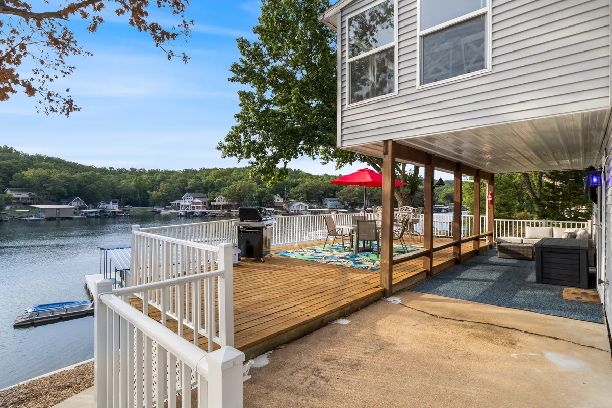 A spacious deck area with chairs and a table shaded by a red umbrella, a grill for barbecuing, and an inviting view of the lake in the background. The setup suggests a perfect spot for social gatherings, outdoor dining, or simply relaxing by the water while enjoying the surrounding scenery.