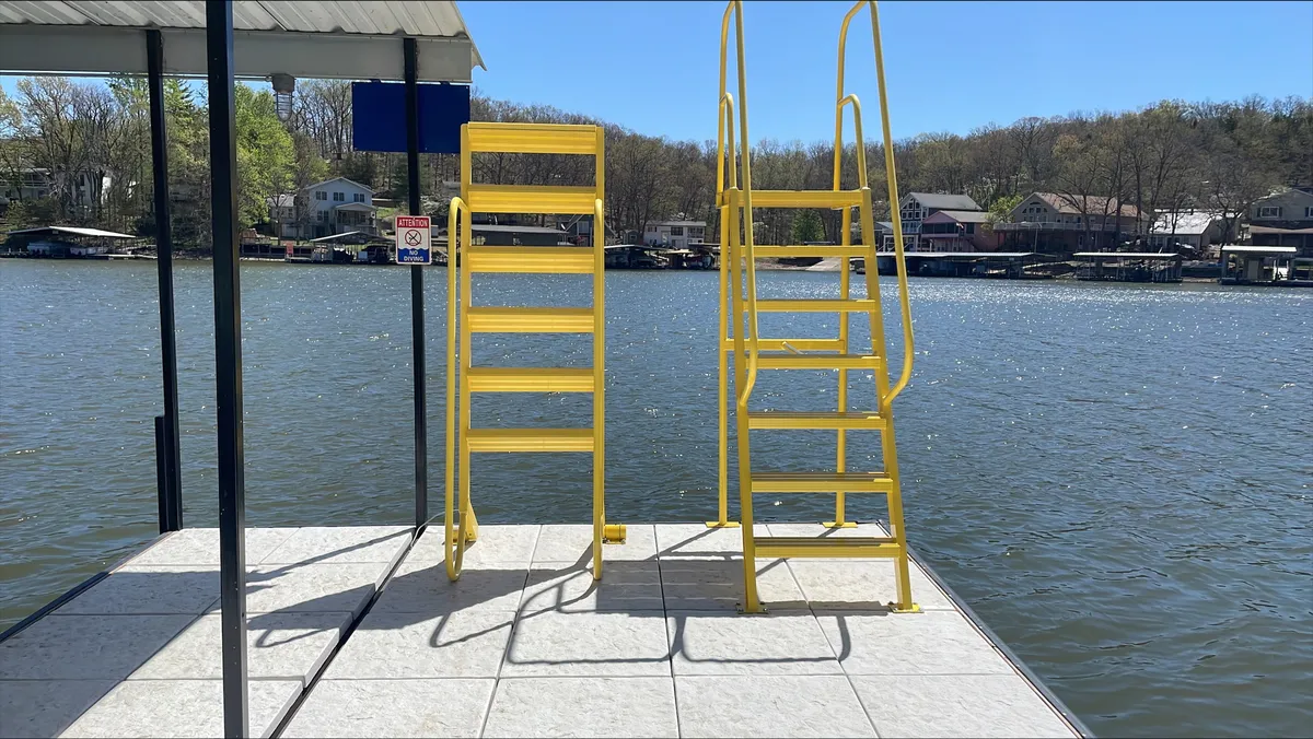 This exterior shot closely frames yellow ladders descending into calm water, with the dock just out of frame and distant houses across the lake. The inviting water surface and easy entry points underscore the playful, accessible water activities mentioned in the section.