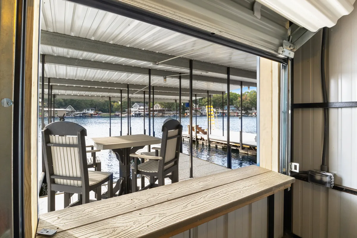 This image captures an inviting outdoor seating area with a table and chairs overlooking the water, a dock extending into the lake, and neighboring houses nestled among trees. The tranquil water view is central, creating a peaceful lakeside atmosphere ideal for relaxation or social gathering.