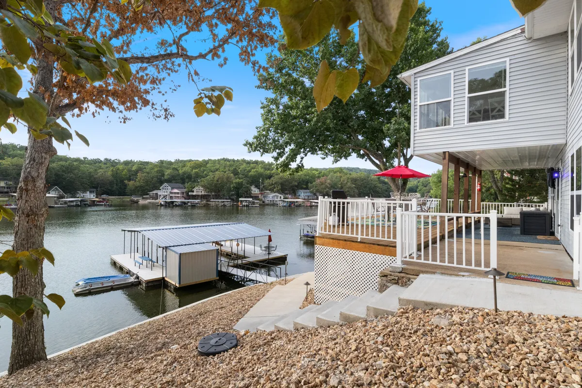 This exterior image showcases a serene deck area overlooking the lake, with a clear view of the water, a boat dock, and surrounding trees. The seating area on the deck is positioned to enjoy the outdoors and direct access to the lakeshore.