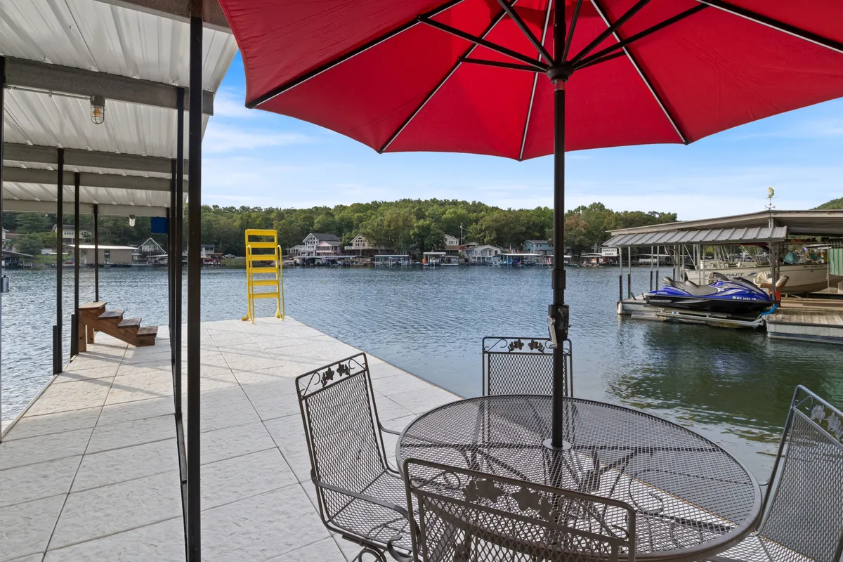 This exterior image features an outdoor dining table under a red umbrella with a sweeping view across the lake, dotted with boats in the distance. The setting radiates a relaxed, social vibe—perfect for watching the lake activity, enjoying meals, or supervising kids as they play nearby.