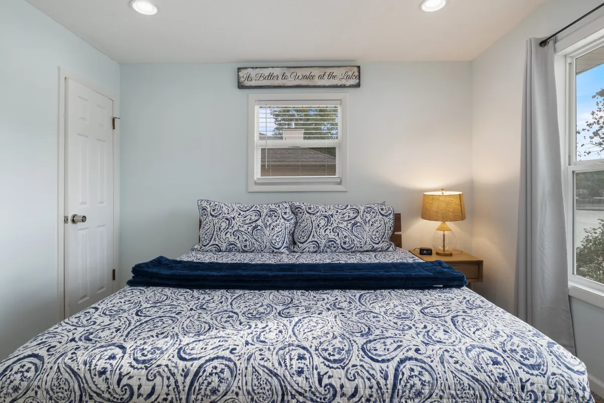 A tranquil bedroom with a patterned bedspread, a window drawing in soft natural light, a nightstand with lamp, and a decorative sign above the bed. The space feels intimate and welcoming, suitable for guests of all ages—including those with pets.